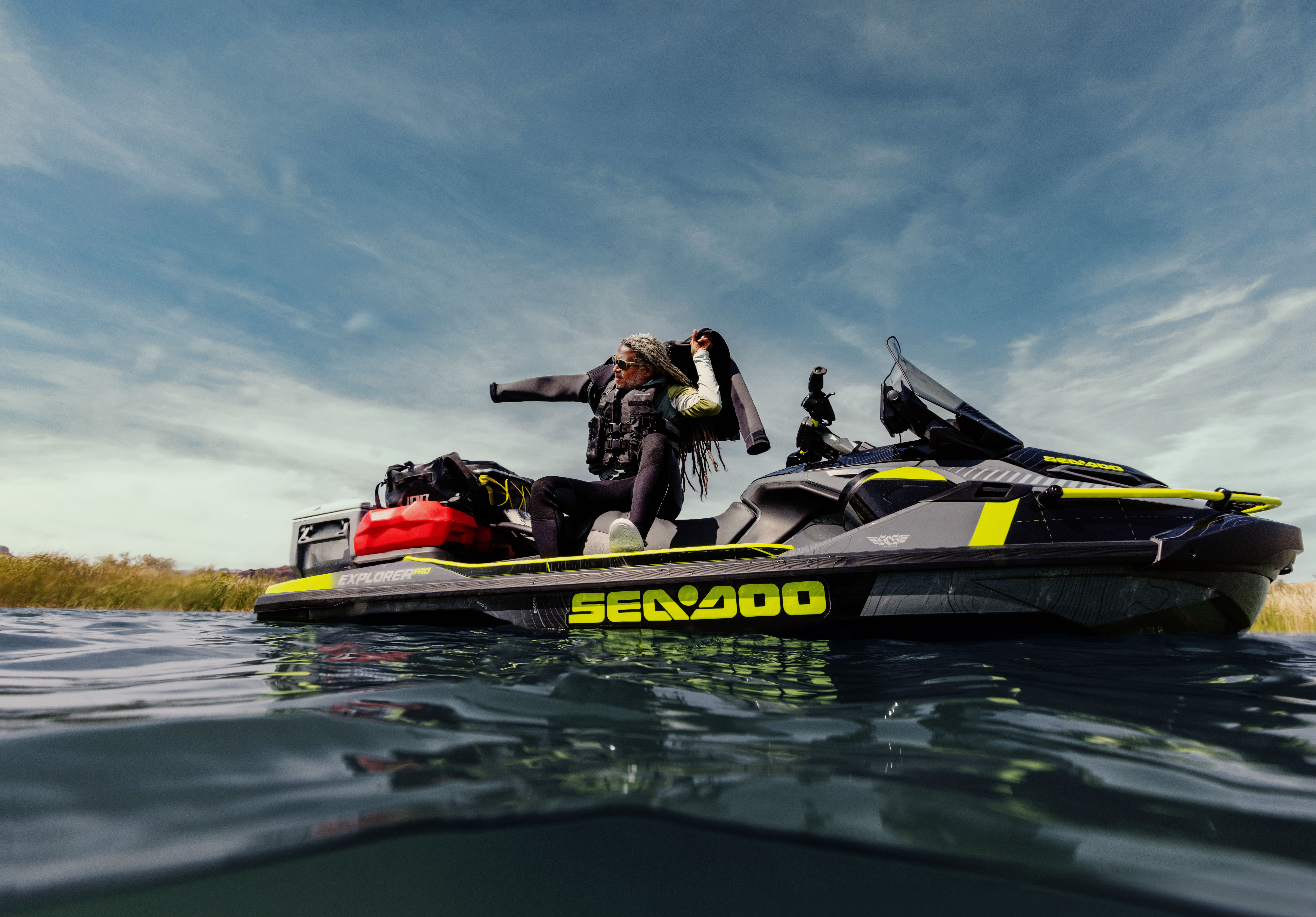 Pitkähiuksinen, päivettynyt mies istumassa Sea-Doo Explorer vesijetin päällä ja laittamassa takkia päälle. Keli on kirkas ja miehellä on mukanaan LinQ lisävarusteita kuten ylimääräinen polttoainetankki.
