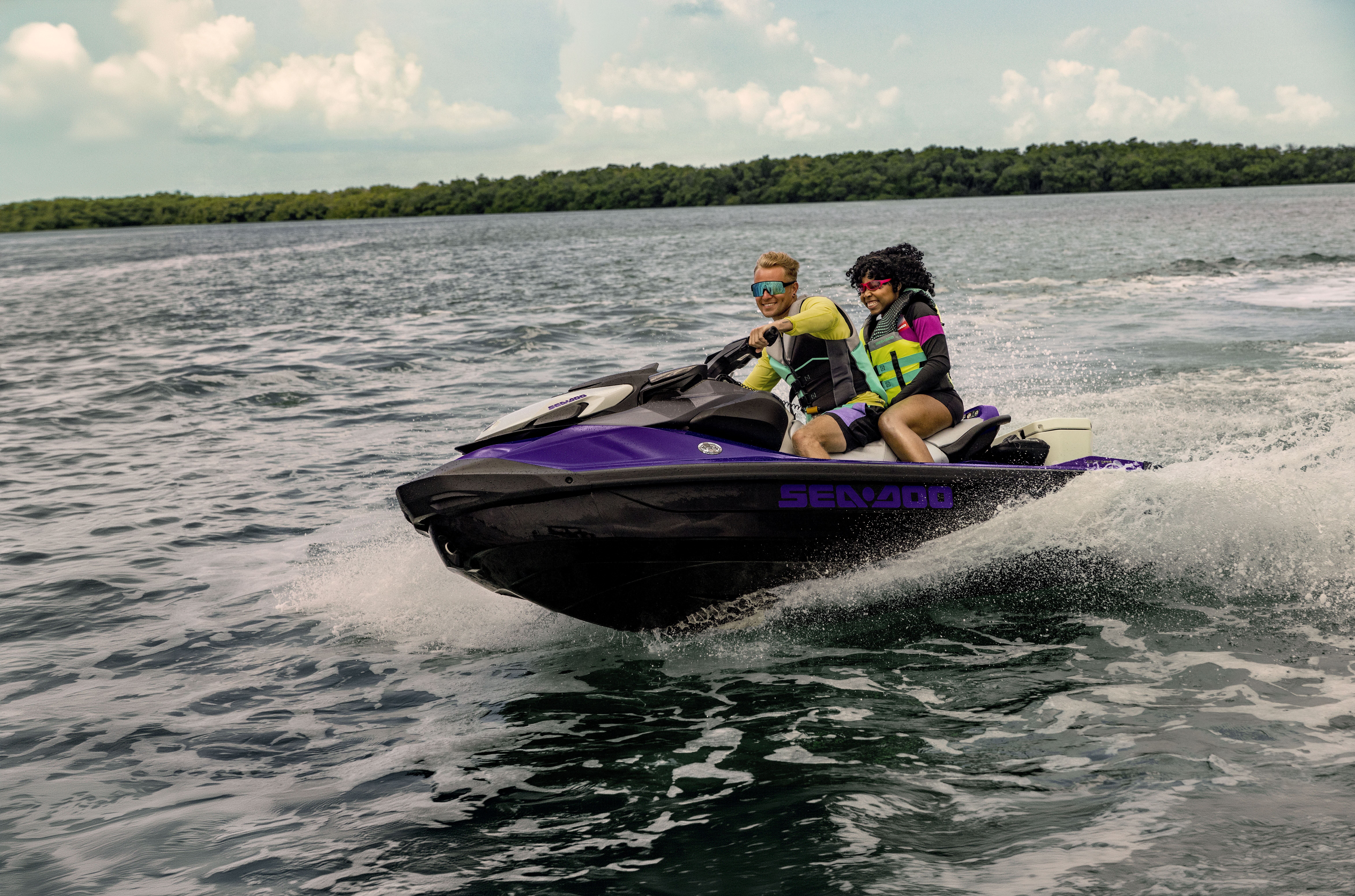 Mies ja nainen ajamassa violetilla Sea-Doo vesijetillä. Mies ja nainen hymyilevät, vesi roiskuu ja vesijetti on hieman kallellaan kaarteessa.
